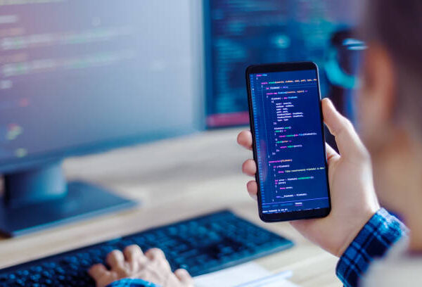 Closeup freelancer hand is holding smartphone with program code. Programmer developer is writing software on multiple computer screens at home office. Devices for working. Geek workplace concept.